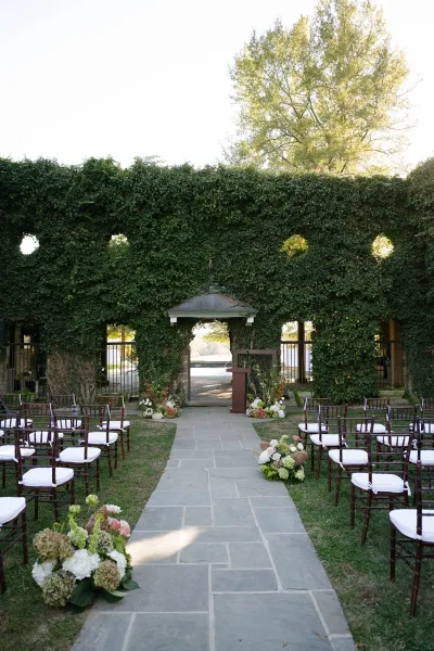 Ceremony aisle design with wood chairs and white cushions framing hydrangea and greenery florals along a stone walkway to an ivy arch doorway