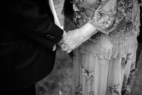 Wedding handholding as bride and groom clasp hands with wedding rings, lace sleeve and suit jacket visible near a white chair on grass