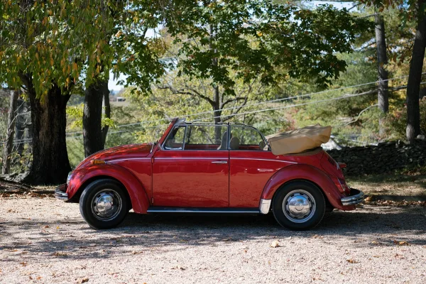 Vintage wedding car, a red convertible with tan soft top and chrome bumpers, parked on a gravel driveway by trees and stone wall