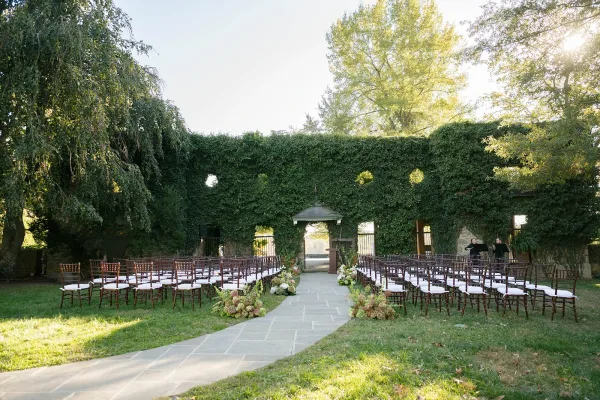 Ceremony setup for an outdoor wedding ceremony with wood chairs and white cushions lining a stone aisle with flowers on a garden lawn