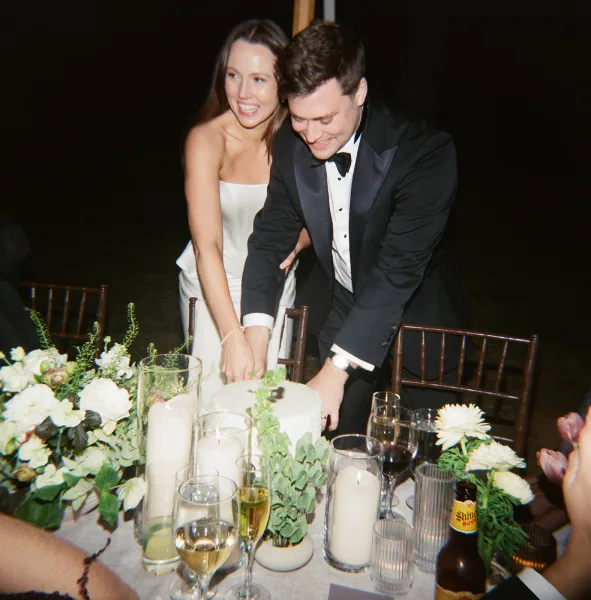 Wedding cake cutting as bride in strapless gown and groom in black tuxedo slice a classic white cake at a candlelit outdoor night table