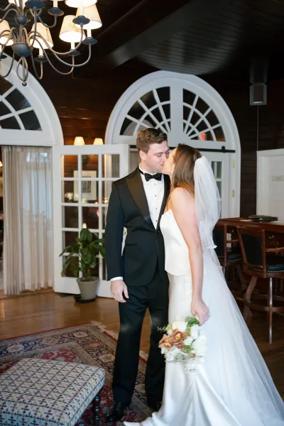 Wedding kiss portrait of bride and groom kissing indoors, bride in veil holding bouquet under chandelier by arched windows and wood walls