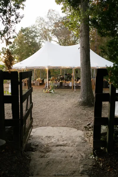 Outdoor wedding tent with white sailcloth canopy and string lights over round reception tables, lanterns, and florals beside trees and a stone path