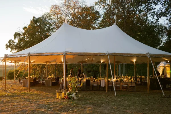 Wedding reception tent with sailcloth wedding tent, farmhouse tables, candles and chandeliers under string lights on a lawn at sunset