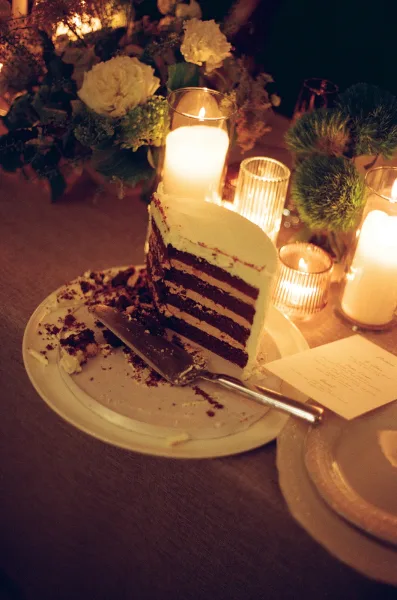Wedding cake with a wedding cake slice on a plate beside a server, surrounded by pillar candles and florals on a dim candlelit reception table