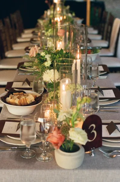 Reception tablescape with wedding table centerpiece, greenery garland, taper candles in glass hurricanes, and place cards in a dim reception space