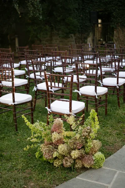 Ceremony seating with brown chiavari chairs and white cushions arranged in rows along a stone walkway on a garden lawn with hydrangeas