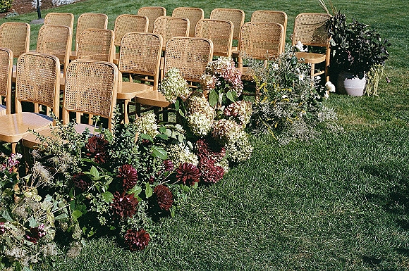Ceremony aisle decor with rattan wedding chairs lined in rows, accented by low floral arrangements and greenery on a grass lawn