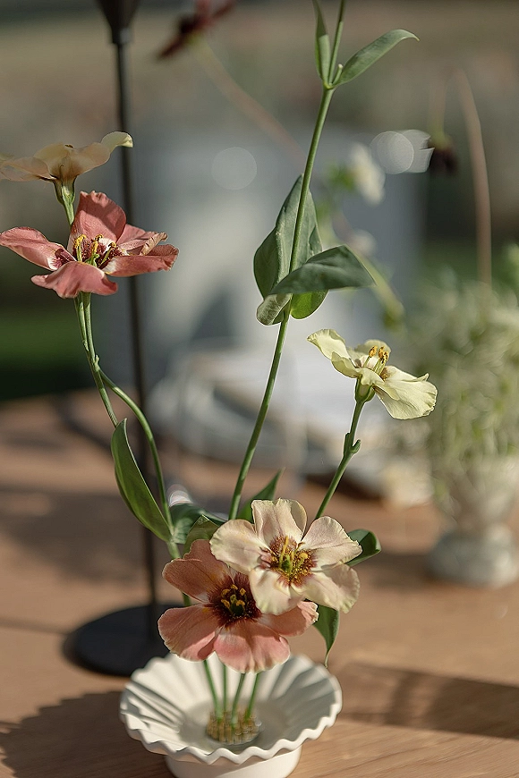 Wedding centerpiece with wildflowers in a scalloped ceramic bud vase on a wooden table, set against blurred reception bokeh lights and greenery