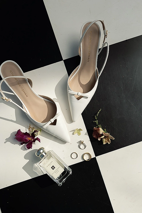 Bridal shoes, white slingback heels on a black and white checkered floor with wedding rings and scattered flower petals nearby
