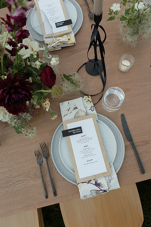 Reception tablescape with wedding place setting on a wood dining table, gray plates, floral napkins, menu cards, taper candles, and centerpiece on grass lawn