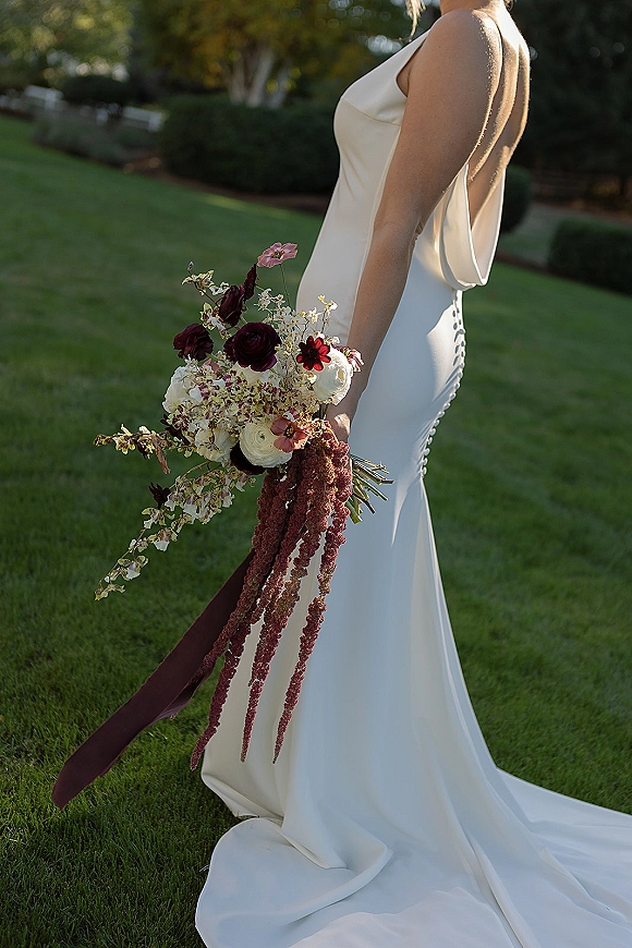 Bridal portrait of a bride in a backless wedding dress with buttoned train, holding a cascading bouquet with burgundy ribbon in a garden lawn