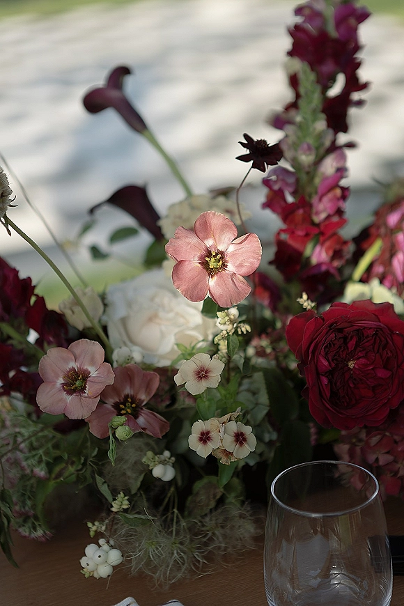 Wedding centerpiece with burgundy wedding centerpiece blooms—blush flowers, white roses, calla lilies, and greenery beside a wine glass outdoors