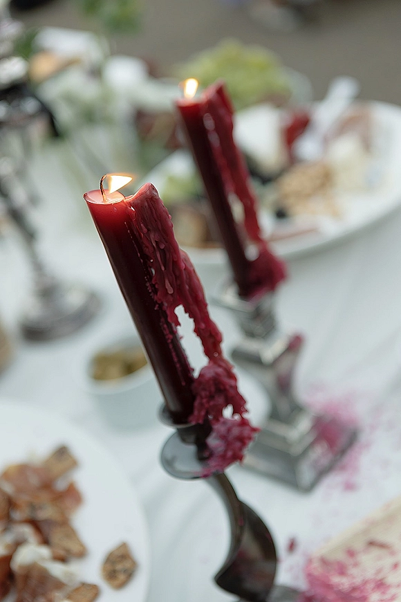 Wedding candle decor with burgundy taper candles in metal candlesticks, wax dripping over a white tablecloth beside plates and glassware