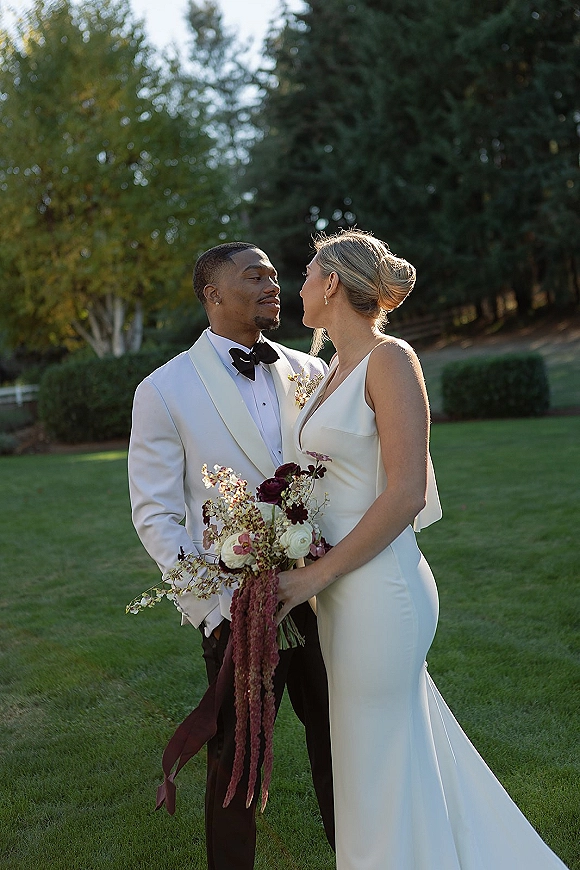 Couple portrait of bride and groom portrait looking at each other, bride holding a burgundy and ivory bouquet on a garden lawn with trees