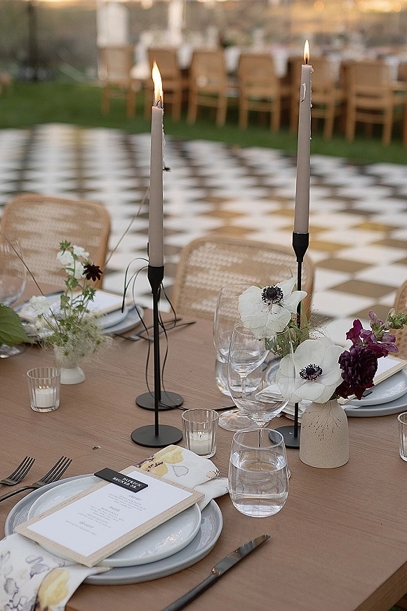 Reception tablescape with taper candle centerpiece, anemone and burgundy blooms on linen tablecloth, string lights and checkered dance floor behind