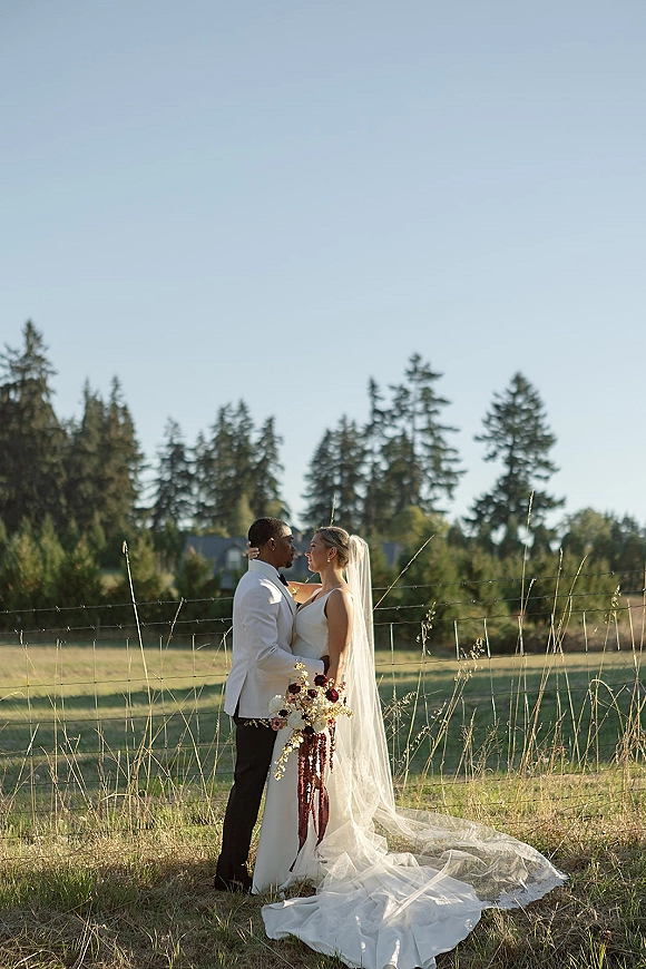 Couple portrait of bride and groom embracing, her long veil trailing as she holds a cascading bouquet in a meadow field by a wire fence