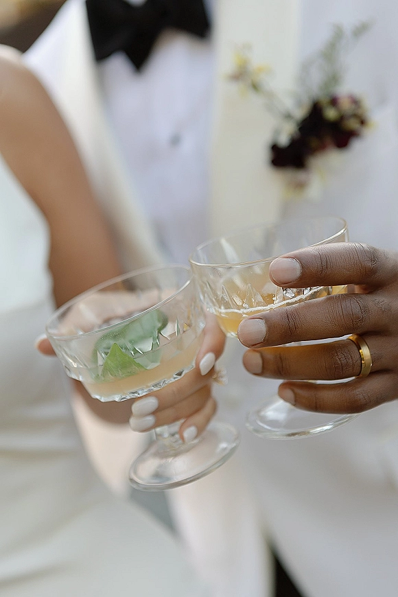 Wedding toast with champagne coupe glasses clinking, mint-garnished cocktails, wedding ring and bow tie details against a white wall