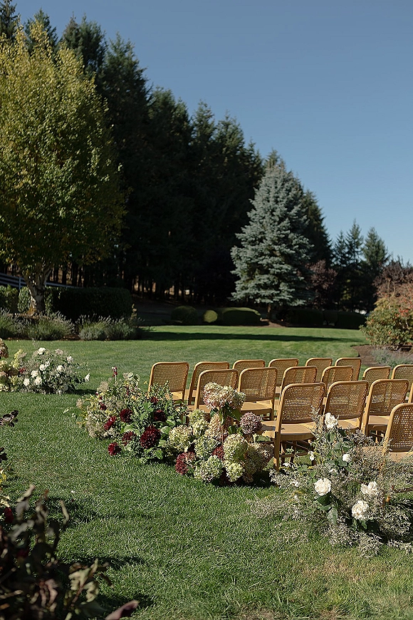 Ceremony seating with outdoor ceremony chairs lined on a green lawn, paired with low rose and hydrangea aisle flowers under blue sky