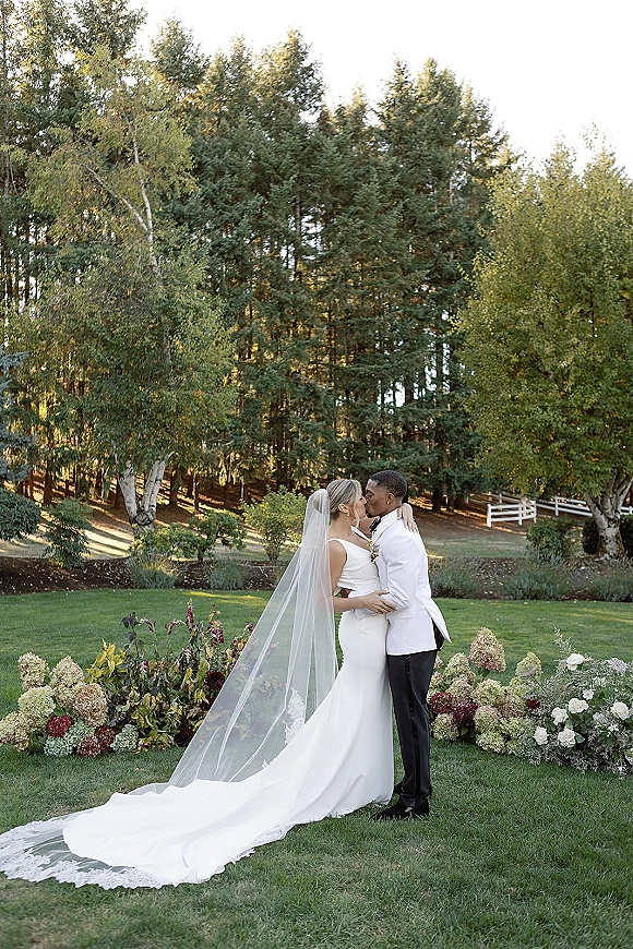 Wedding kiss portrait of bride and groom kissing from behind, bride’s long veil train flowing on a garden lawn by a white fence