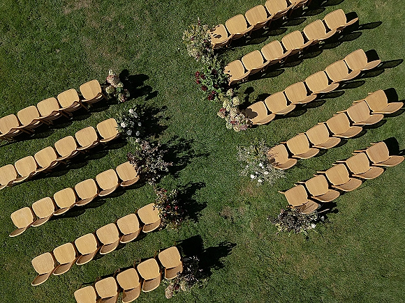 Ceremony seating layout with curved aisle and wood chairs, framed by floral arrangements and greenery on a grass lawn outdoors
