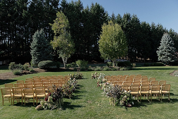 Outdoor ceremony setup with wood ceremony chairs lining a floral aisle, greenery and ground arrangements on a lawn by forest edge under blue sky