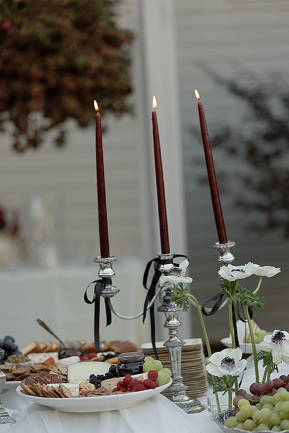 Wedding grazing table with wedding charcuterie board, cheese and fruit, taper candles, and silver candelabra on a white tablecloth outside a white building