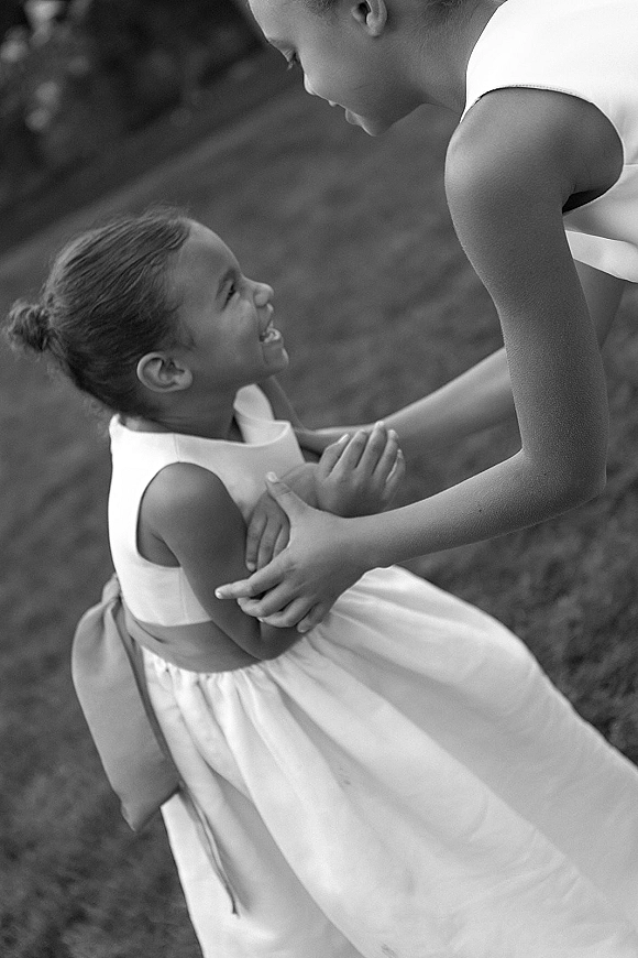 Flower girl moment in a flower girl dress with a sash, hair bun, and bow, standing on a grass lawn during the wedding day