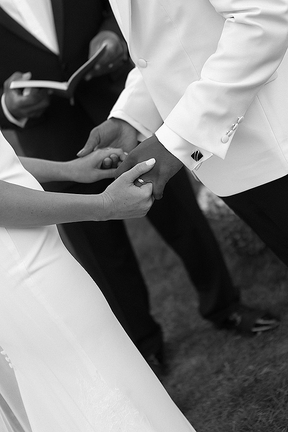 Ceremony moment as bride and groom hold hands during vows, wedding rings visible on groom in white suit jacket with cufflinks, guests seated behind