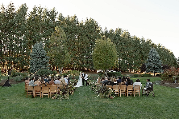 Outdoor wedding ceremony with bride and groom holding hands at the altar, guests in wood chairs along a floral aisle on a lawn with trees