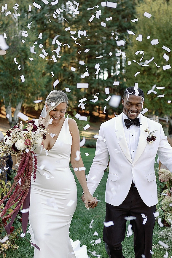 Recessional moment with wedding confetti toss as bride and groom walk hand in hand on a garden lawn, guests behind, bouquet and veil flowing
