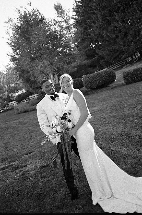 Couple portrait in black and white wedding portrait style, bride with long train holding bouquet, groom in white tux, on lawn by trees and fence