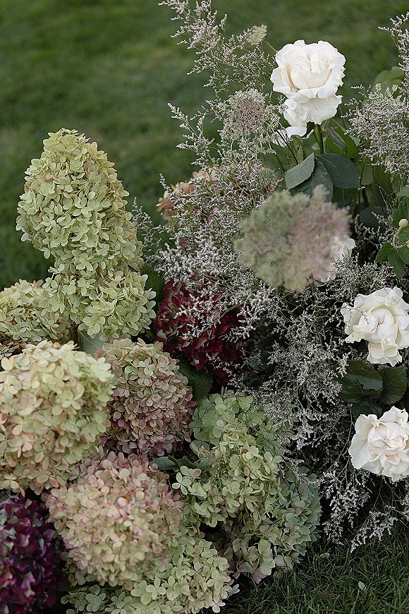 Wedding floral arrangement with hydrangea wedding flowers, white roses, and airy greenery set low on a grass lawn for ceremony decor