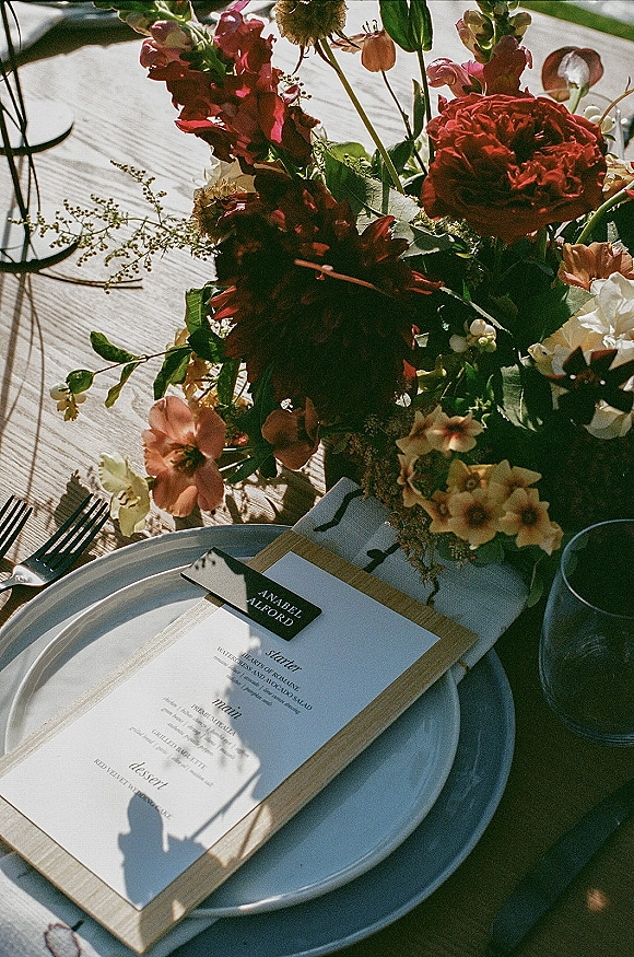 Reception tablescape with a floral centerpiece, white plates and forks, and a menu card on a wooden clipboard in outdoor light on a wood table
