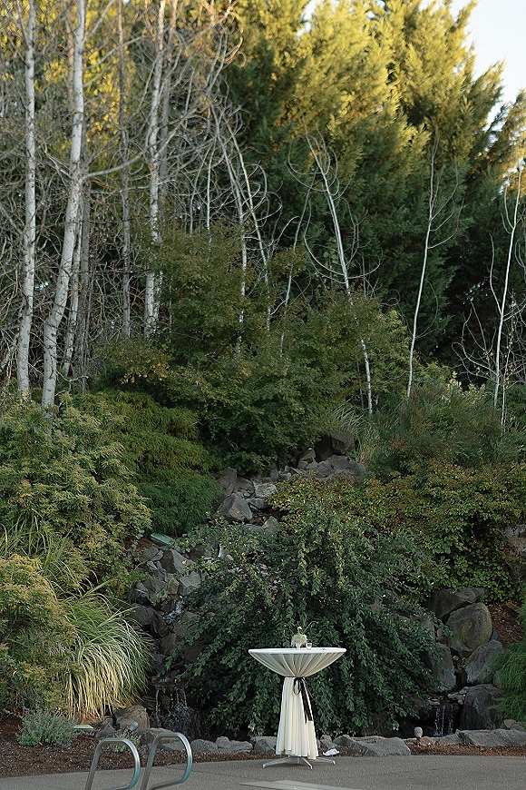 Cocktail hour setup with a high top cocktail table decor in white linen and black ribbon, bud vase and votive candles by a small waterfall