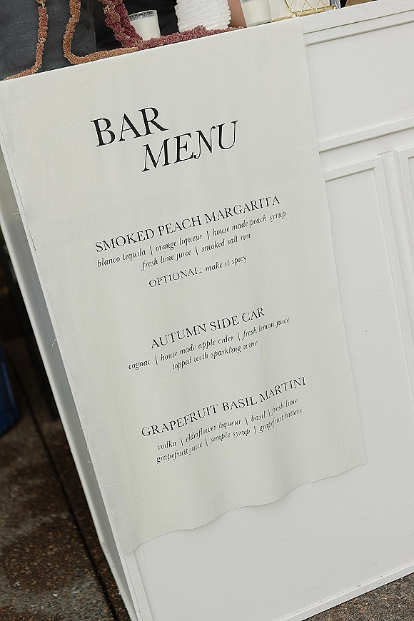 Bar menu sign with wedding bar menu details in classic black and white typography, displayed in an acrylic holder on stone floor by white paneling