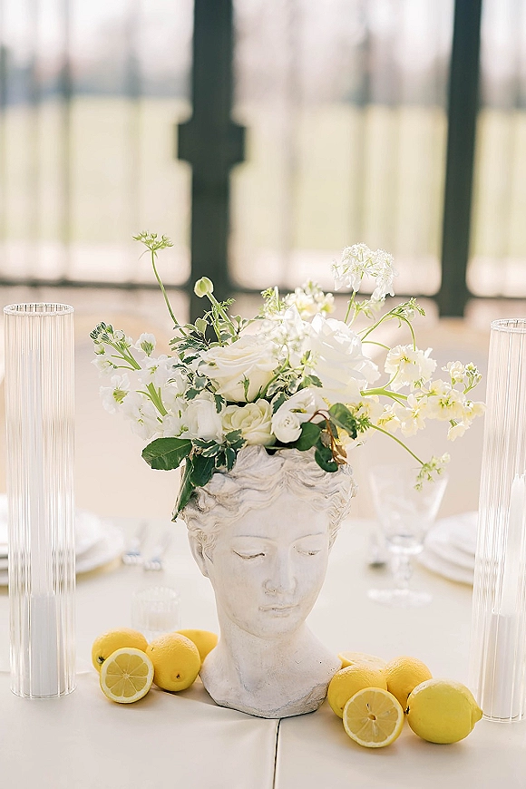 Wedding centerpiece with white flowers and greenery in a classical bust vase, lemons and ribbed glass vases on a table by large windows