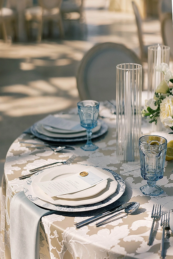 Reception tablescape with blue and white tablescape details, patterned cloth, white tapers in glass vases, floral center and lemons on sunlit patio