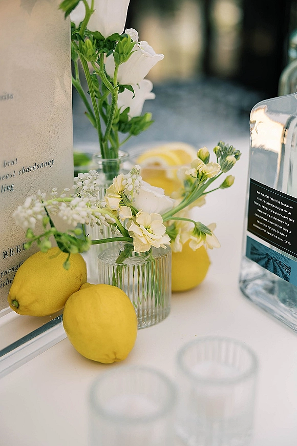 Cocktail table decor with lemon wedding decor, clear bud vases of white and yellow flowers, striped glass, and drink menu card outdoors