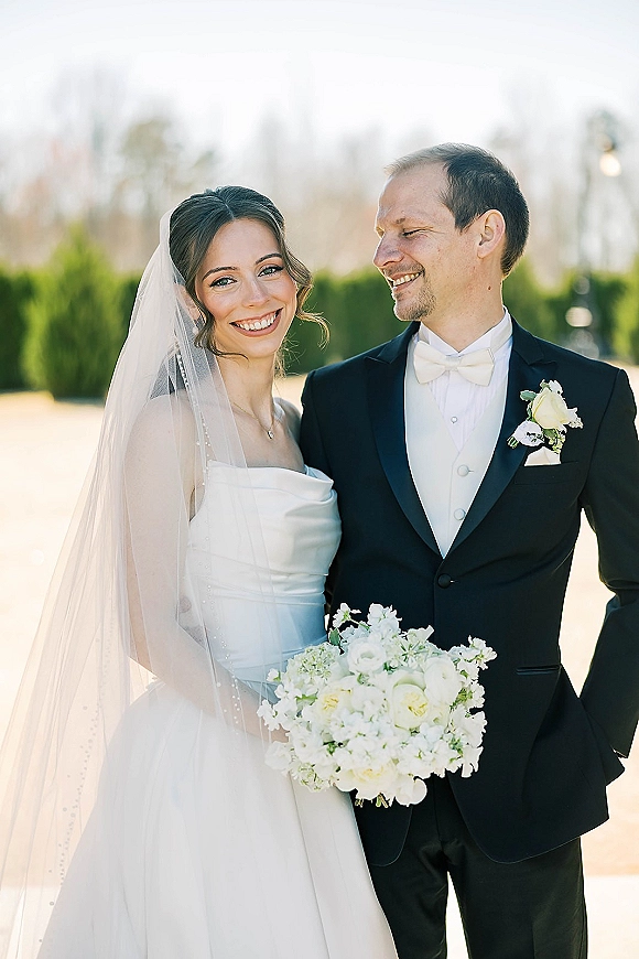 Couple portrait of bride smiling at camera as groom looks at her, her veil and white bouquet bright against sunlit trees outdoors