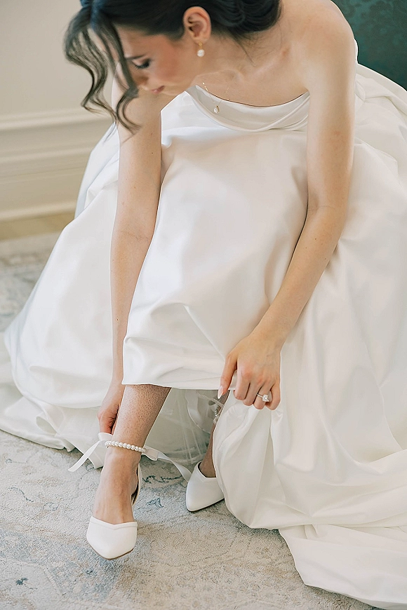 Bridal getting ready detail with bride putting on shoes, strapless satin wedding dress draped, pearl garter and ring on a patterned rug