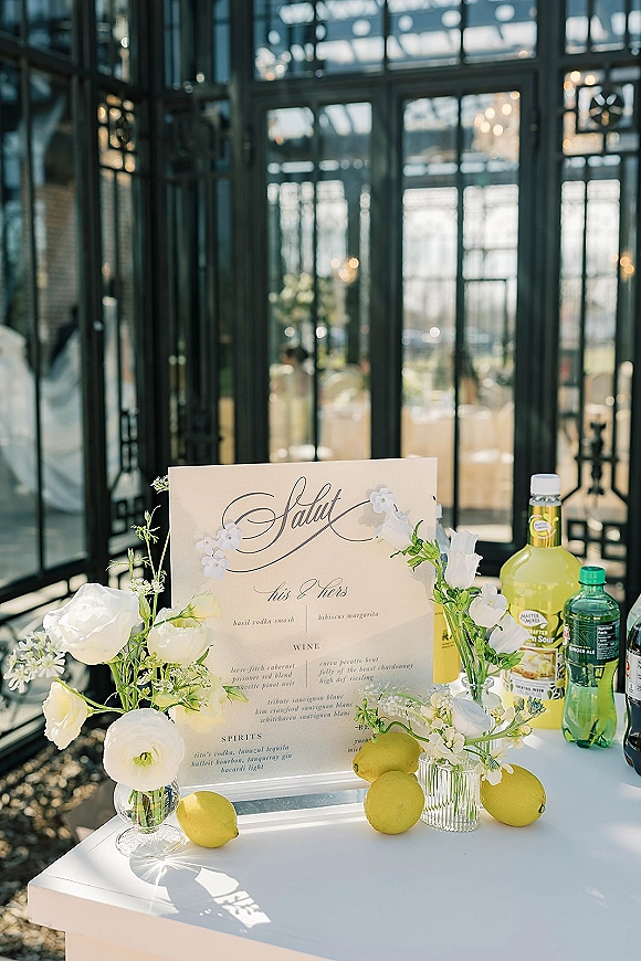 Wedding bar menu with a signature cocktail menu sign in an acrylic holder, accented by white flowers and lemons in a sunlit greenhouse setting