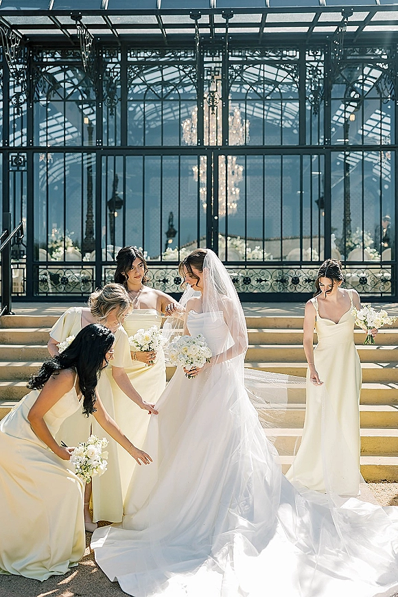 Bride with bridesmaids as bridesmaids adjusting dress and veil, holding white rose bouquets on stone steps in a glass conservatory chandelier backdrop