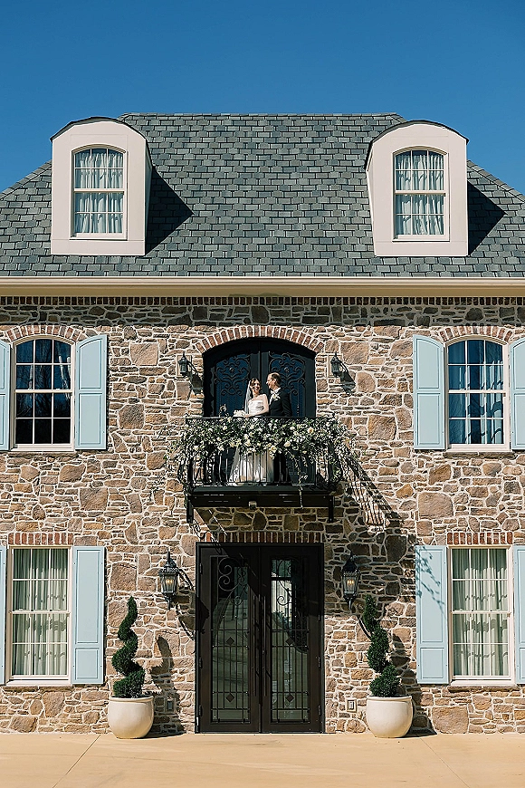 Couple portrait of bride and groom balcony embrace in formal attire with floral garland, set against a stone facade with blue shutters