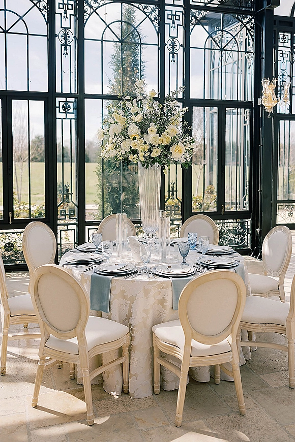 Reception tablescape with a round table, tall fluted vase of white roses and greenery, dusty blue runner, set under a chandelier in a glass conservatory