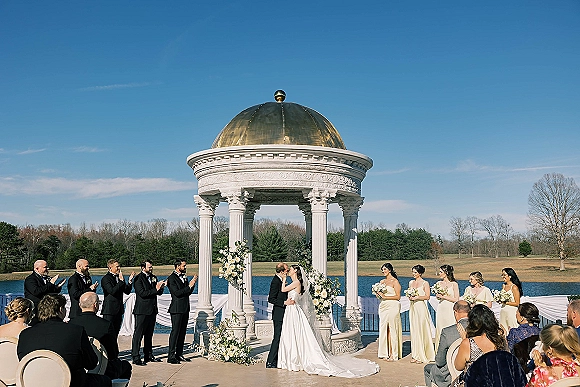 Ceremony kiss beneath a flowered wedding arch as the couple stands by a lake, bride’s veil trailing, guests and bridesmaids behind