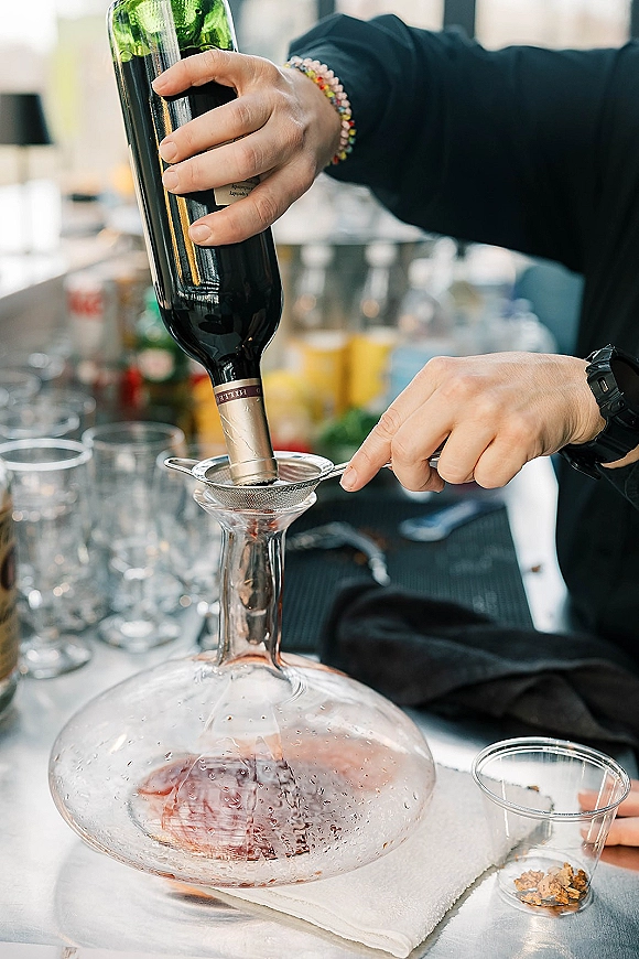Wine decanting at a reception bar setup, hands pouring from bottle through mesh strainer into decanter on a bar counter with blurred bottles behind