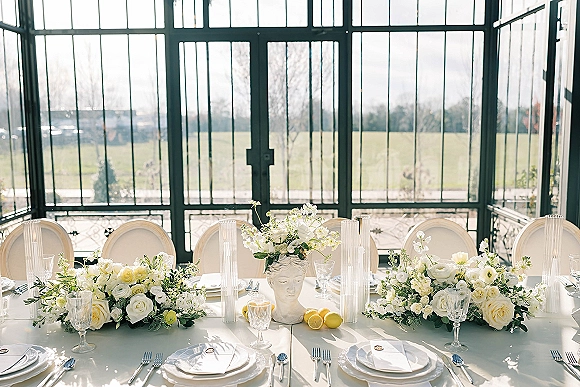 Reception tablescape with white floral centerpieces and greenery garland, taper candles, lemons, and crystal stemware in a glass conservatory