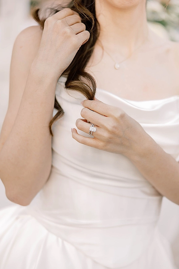 Engagement ring close-up showing a diamond engagement ring and wedding band stack on a bride’s hand beside a pearl pendant and satin dress neckline