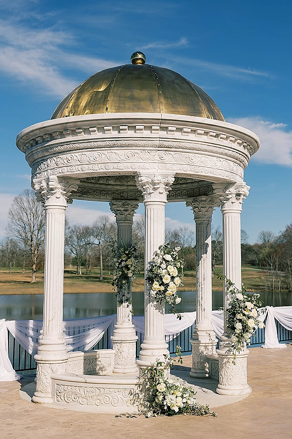 Wedding ceremony altar with white floral arrangements and greenery garlands, draped on a round gazebo with gold dome by a lake under blue sky